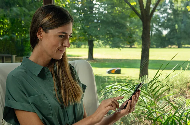 A woman that is controlling  her autonomous robot mower through her smartphone